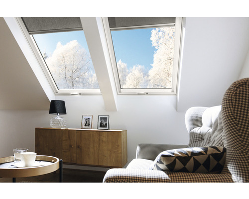 Chambre mansardée lumineuse avec fenêtres de toit et stores, meublée avec une commode, une lampe et un fauteuil