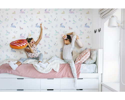 Deux filles jouent sur un lit devant un papier peint à motifs de licorne.