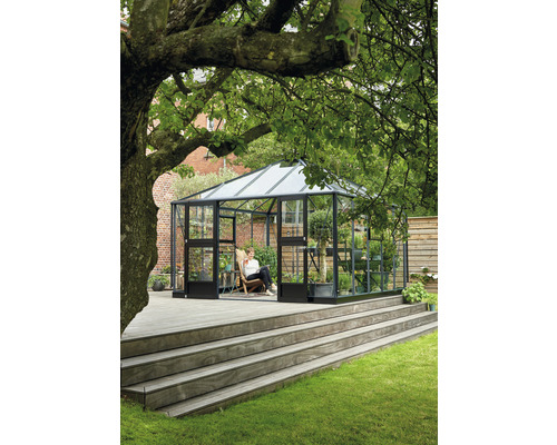 Ein Gewächshaus aus Glas und Metall steht auf einer Holzterrasse in einem Garten.