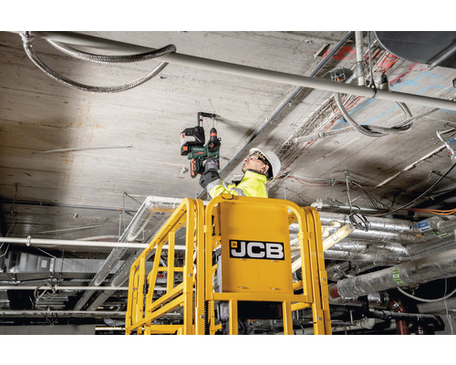 Un homme sur une plateforme de travail avec le logo JCB perce un plafond en béton avec un perforateur sans fil.