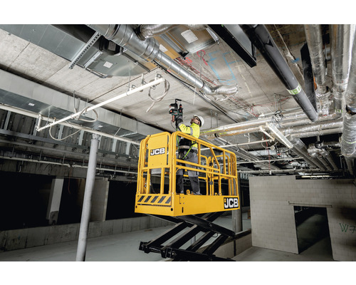 Un ouvrier sur une plateforme élévatrice à ciseaux jaune travaille au plafond d''une pièce non finie avec des tuyaux et des conduits apparents.