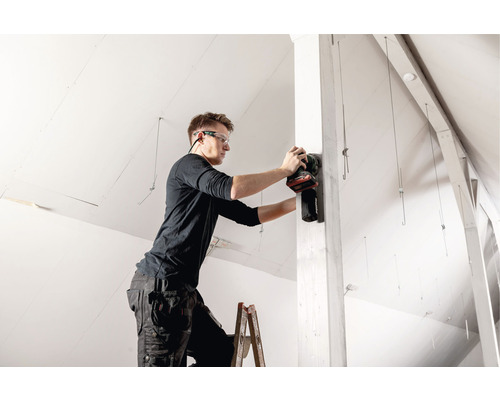 Artisan sur une échelle ponçant un poteau en bois avec une ponceuse électrique dans un grenier.