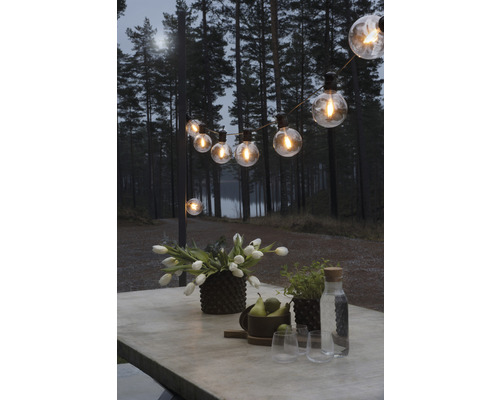Table décorée avec des fleurs, des fruits et des verres sous une guirlande lumineuse à l''extérieur.
