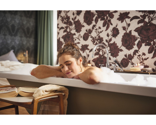 Frau entspannt in der Badewanne mit Schaum vor einer floralen Tapete