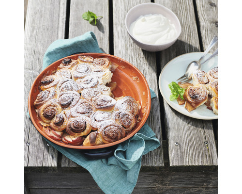 Apfel-Zimtschnecken in einer Auflaufform mit Puderzucker bestreut, dazu ein Schälchen Creme und ein Teller mit Zimtschnecken
