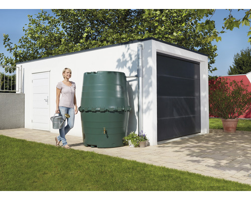 Eine Frau steht mit einer Gießkanne neben einer Regentonne vor einer Garage.