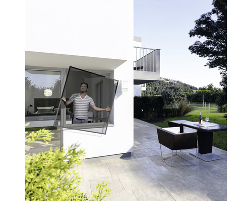 Mann montiert einen Insektenschutz an einem Fenster eines Hauses mit Terrasse und Gartenmöbeln.