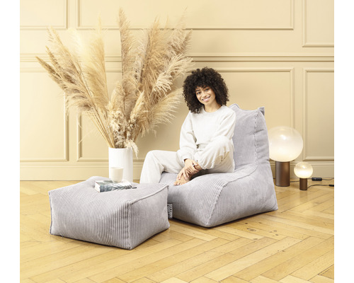 Une femme est assise dans une pièce sur un pouf en velours côtelé gris avec un tabouret assorti.
