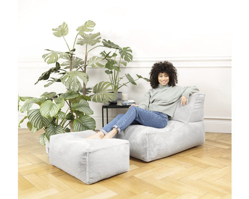 Une femme est assise dans un salon lumineux sur un pouf gris avec un repose-pieds assorti