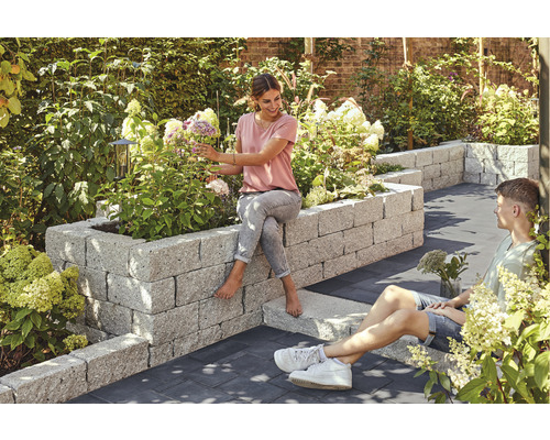 Jardin avec parterre surélevé en pierre, planté de fleurs, sur lequel une femme est assise, tandis qu''un homme est assis par terre à côté