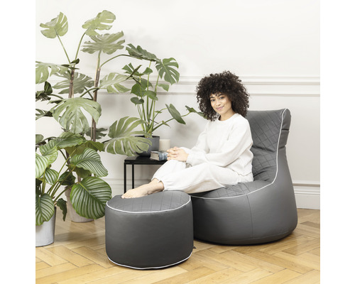 Une femme est assise dans un fauteuil gris avec repose-pieds dans une pièce lumineuse.