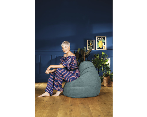 Frau sitzt in einem Zimmer auf einem Sitzsack, umgeben von Zimmerpflanzen und Bildern.