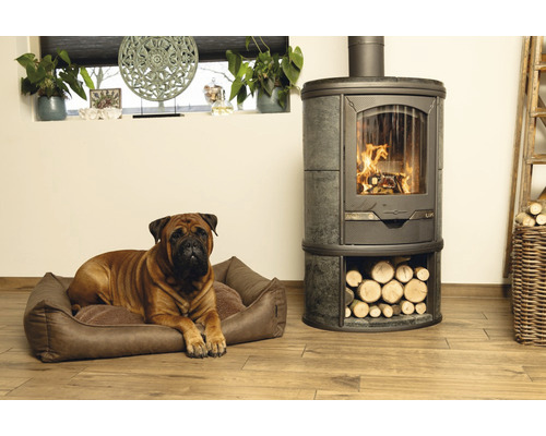 Bullmastiff liegt in einem Hundebett neben einem Kaminofen mit Holzfach