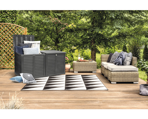 Gartenszene mit grauem Aufbewahrungsschrank, Rattansofa und schwarz-weissem Teppich auf einer Holzterrasse.