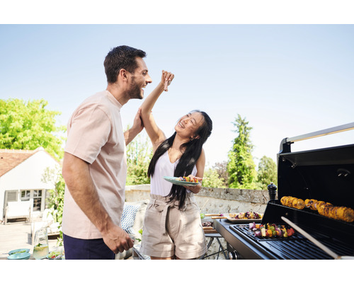 Deux personnes font des grillades sur une terrasse avec un barbecue à gaz. Des brochettes et des épis de maïs sont posés sur le gril.