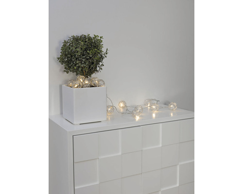 Guirlande lumineuse décorative avec des lampes sphériques sur une commode blanche à côté d''une plante en pot.