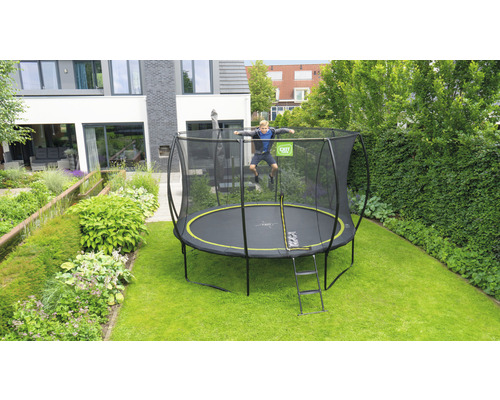 Garçon sautant sur un trampoline de jardin avec filet de sécurité et échelle devant une maison.