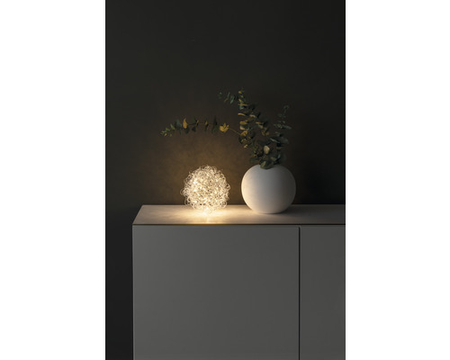 Dekoratives Arrangement mit einer leuchtenden Drahtkugel, einer Vase und Zweigen auf einem Schrank.