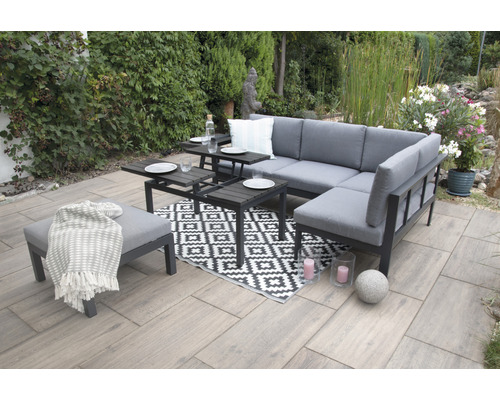 Scène de jardin avec ensemble de salon d''angle gris, tables d''appoint, tabouret et tapis à motifs sur une terrasse.