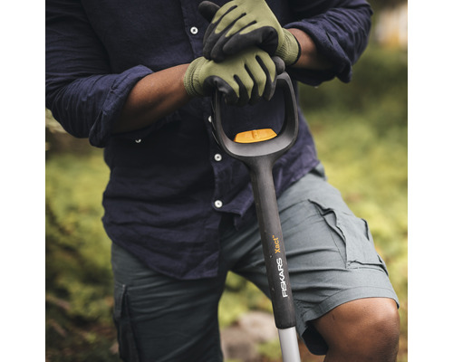 Une personne portant des gants de jardinage tient une bêche de la marque Fiskars.