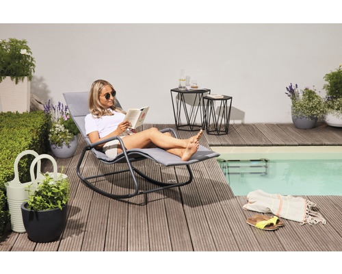 Szene im Außenbereich mit Frau, die ein Buch auf einer Gartenliege am Pool liest. Zwei Beistelltische, Pflanzen und Dekorationen vervollständigen das Bild.