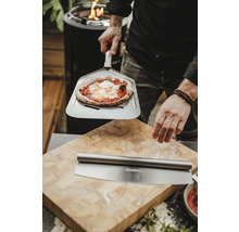 Un homme tient une pizza sur une pelle à pizza devant une planche à découper avec un coupe-pizza
