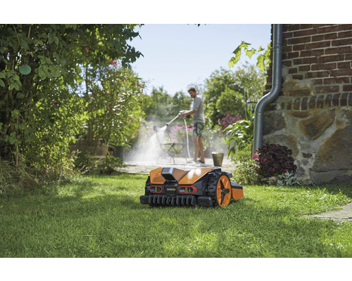 Un robot tondeuse à gazon est posé sur une pelouse dans le jardin. À l''arrière-plan, on aperçoit une personne avec un tuyau d''arrosage.