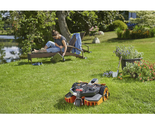 Un robot tondeuse à gazon se trouve dans le jardin, tandis qu''une femme se détend sur une chaise longue.