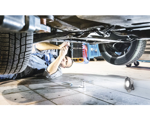 Un mécanicien est allongé sous une voiture et travaille dessus, éclairé par une lampe de travail.