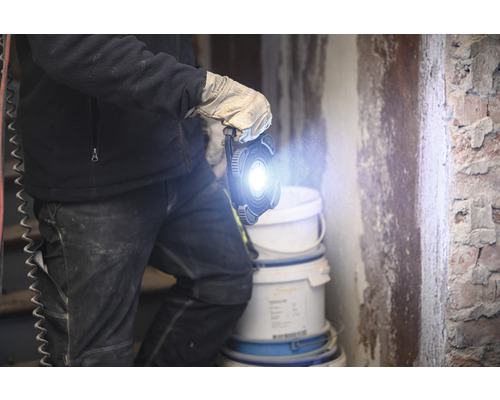 Une personne gantée tient une lampe de travail à LED dans une pièce avec des seaux de peinture.