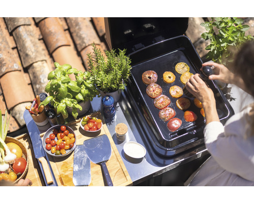 Une personne prépare des tranches de tomates sur un gril, à côté desquelles se trouvent des herbes fraîches et des ingrédients.