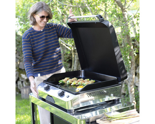 Femme préparant des aliments sur une plaque de cuisson à gaz en acier inoxydable avec des grillades et des légumes.