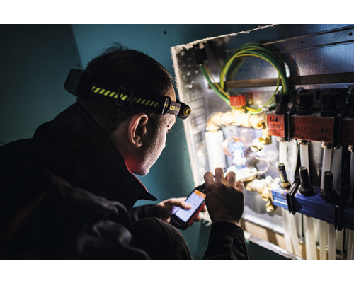 Un homme avec une lampe frontale vérifie un système de chauffage avec un smartphone.