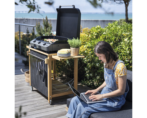 Scène avec une femme assise devant un ordinateur portable et un barbecue sur un chariot en bois à l''extérieur.