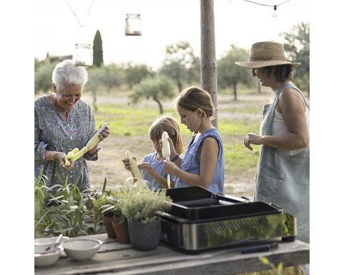 Scène en extérieur avec une famille épluchant des épis de maïs et utilisant un grill de table.