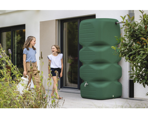 Szene mit zwei Mädchen und einem grünen Regenwassertank vor einem Haus.