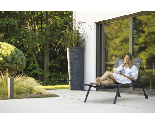 Frau liest Buch neben Regentonne im Garten.