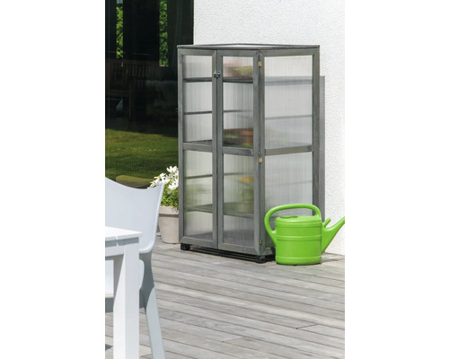 Armoire en bois grise avec étagères et panneaux transparents sur une terrasse à côté d''un arrosoir vert.