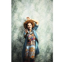 Eine Frau mit Afrofrisur und gemustertem Kleid vor einer Tapete mit Palmenmuster