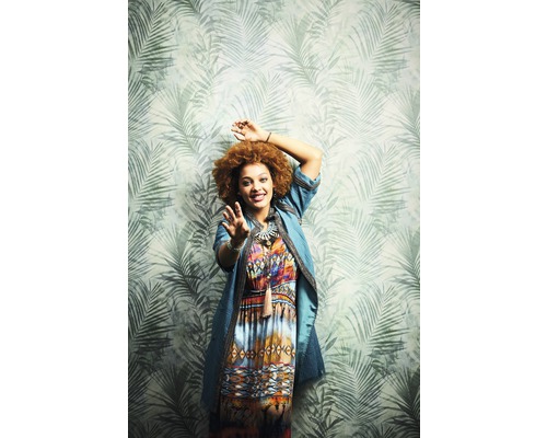 Eine Frau mit Afrofrisur und gemustertem Kleid vor einer Tapete mit Palmenmuster
