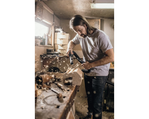 Un homme nettoie un atelier de menuiserie avec un aspirateur, tandis que des copeaux de bois flottent dans l''air.
