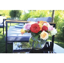 Ein Arrangement aus Rosen in einer Glasvase auf einem Gartentisch mit Gartenmöbeln im Hintergrund.