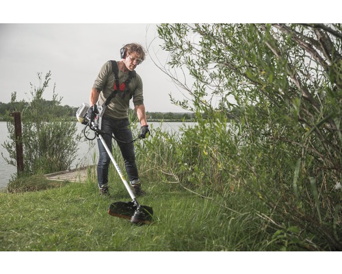 Mann bedient Rasentrimmer am Seeufer mit Schutzbrille und Kopfhörern