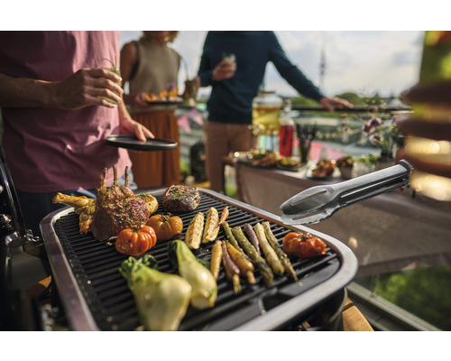 Grillszene mit Fleisch, Gemüse und Grillzange auf einem Grill, umgeben von feiernden Menschen.