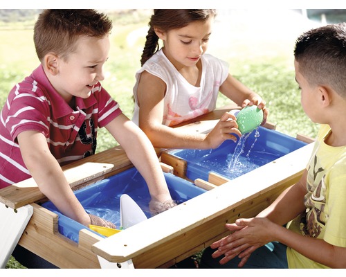 Trois enfants jouent à une table en bois avec du sable et de l''eau et des récipients bleus à l''extérieur.