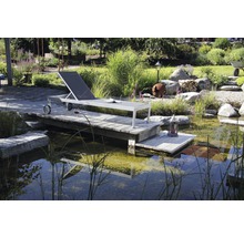 Scène avec une chaise longue en métal sur une plateforme en bois au bord d''un étang dans un jardin.