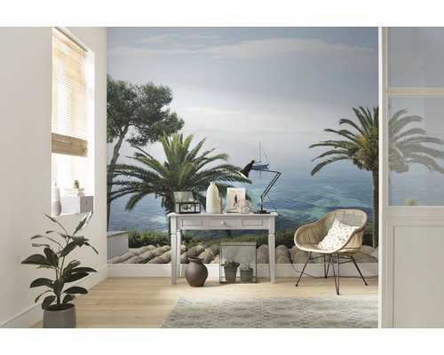 Espace de vie avec un papier peint photo représentant un paysage côtier avec des palmiers et un voilier, meublé d''un bureau et d''un fauteuil en rotin.