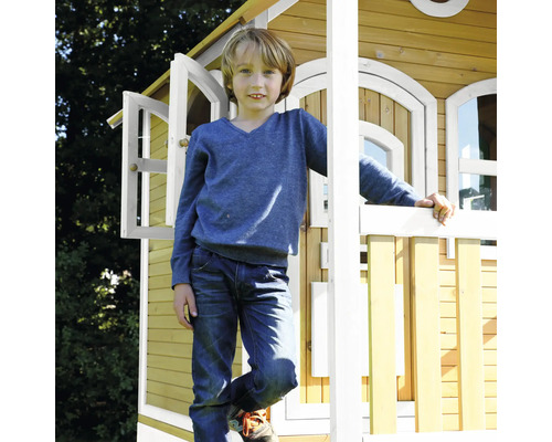 Garçon devant une cabane de jeu en bois avec fenêtres ouvertes.