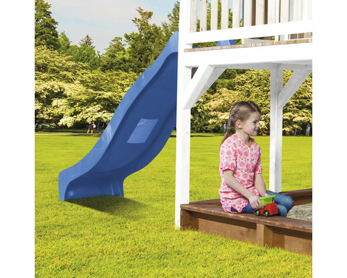 Kind sitzt mit Spielzeug auf einer Sandkiste neben einer blauen Rutsche auf einem Spielplatz