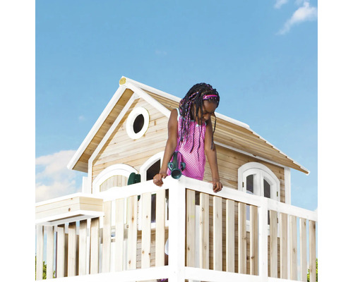 Un enfant se tient sur une cabane en bois avec une balustrade et une petite maison avec des fenêtres sous un ciel bleu.